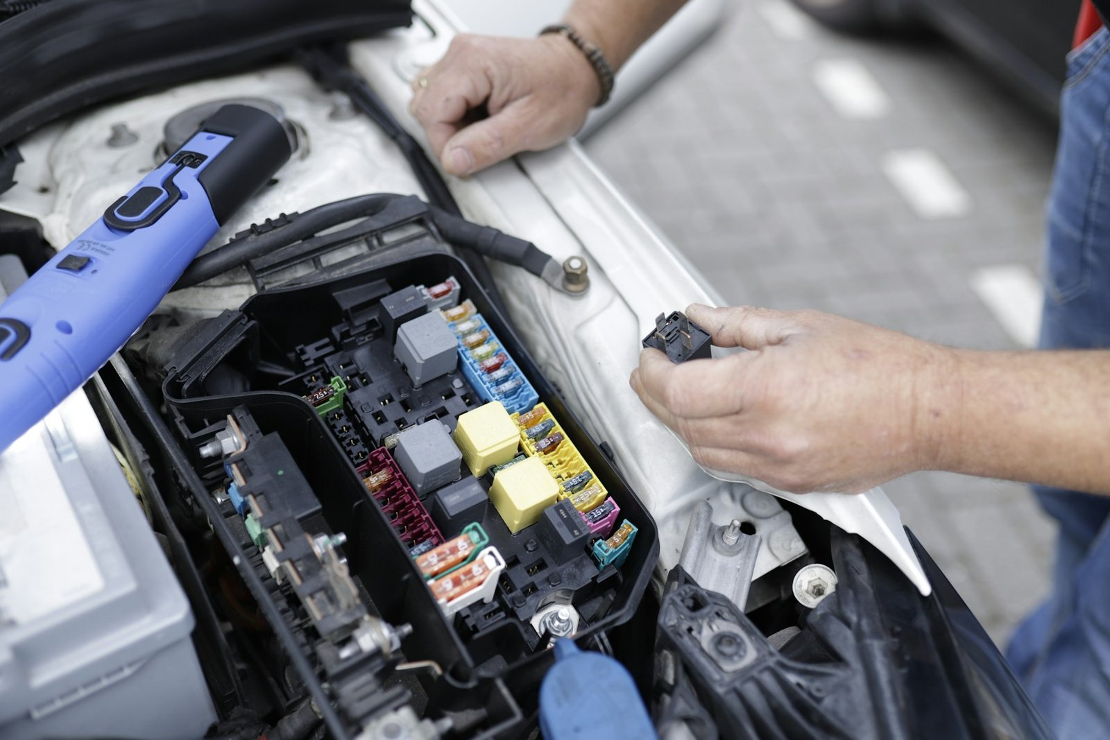 Hands holding a relay beside the open fuse and relay box under the hood of a car